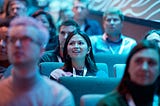 The image shows an audience seated in what appears to be a conference or event setting. The focus is on a young woman in the center, wearing a lanyard and looking engaged, with a bright and excited expression. She is surrounded by other attendees, some of whom are slightly out of focus. The lighting in the room is predominantly blue, creating a cool-toned ambiance. The background suggests that everyone is attentively watching a presentation or speaker. The setting appears modern and professional