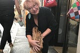 Author Carol Lennox in front of a store for hand painted purses. A cocker spaniel dog is looking up at her as she is smiling at the camera. Solo travel. Italy.