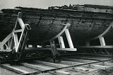 Black and white photograph of a man standing in front of a giant wooden hull