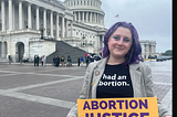 A photo of Lizzie, a White woman with purple hair. She is wearing a T-shirt that reads “had an abortion,” and holding a sign that says “ABORTION JUSTICE NOW. The Capitol building is in the background.