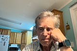 An older man with gray hair sits at a kitchen table, resting his face on his hand and appearing deep in thought or perhaps dejected. He is wearing a plaid shirt and a smartwatch. Behind him are wooden cabinets, a microwave, and a refrigerator with butterfly magnets. The lighting is bright, and the overall mood seems contemplative or sad.