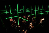 Visitors sit in the Donmar auditorium, 2 metres apart wearing headphones. A grid of green strip lights hangs above them.