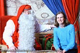 Santa with a long beard in red clothes holding his an arm on a shoulder of a brunette in a blue sweater. Both smiling at camera.