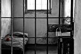 Black-and-white photo of a harsh prison cell on a remote island prison: thick metal bars in the foreground, a narrow bed with a single red rose on the mattress, and a small wooden table with a heavy padlock, illuminated by dim light through a high, barred window.