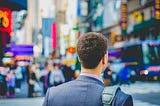 Photo of the upper back-side of a man in a suit, standing and looking into the street.