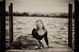 Black and white shot of a woman in a dress on a pier, water behind her.