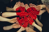 Photo of many light-skinned hands coming together, showing their palms. A large red heart is painted on the palms and hands together, as they’re all placed at the moment. So when the hands separate, the heart will not be a heart shape, but will be various shapes of red paint.