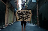 Man in narrow walk way holding a sign that covers his upper body and reads ‘better days.’