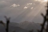 A dark mountain range with light breaking through dark clouds, in the foreground some blurry shapes of branches sticking out from the bottom