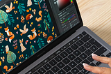 A close-up view of a graphic designer’s hands working on a laptop with design software open, displaying a colorful pattern featuring stylized forest animals and plants on a dark background. The screen shows a vibrant, detailed illustration with various tools and color palettes, indicating the digital creation process of children’s pattern design.