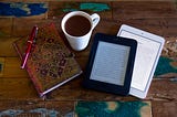 An ebook reader, iPad, a notebook and pen, and a cup of hot chocolate all on a rough wooden table.