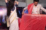 Picture shows a man (who is a Bollywood actor) in a floaral printed shirt and white blazer showing a woman (who is also a Bollywood actor) how to tie a saree; Picture is indicative of a real practice where men saree sellers would often show a woman how the saree would look by tieing it on their own bodies