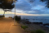 A coastal promenade in france, lit by lampposts in the early morning. The sky is just getting lighter with some clouds scudding the sky. The sea and some rocks and a corner of beach can be seen, with gentle waves coming in and trees lining some of the path