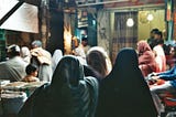 A market in Lahore, Pakistan. Women and men dressed in traditional garb walk through a darkly lit market.