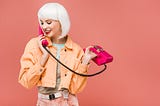 young woman in white-blonde wig wearing an orange jacket talking on a hot pink corded phone on dusty rose background