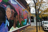 Colorful wall art depicting diverse people lives on a Red Hook building next to black tree with falling yellow leaves