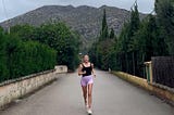 The author runs on the beautiful island of Mallorca, Spain on a open road with a mountain in the horizon and green trees of to both sides.