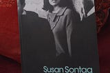 A photograph of the author’s edition of Susan Sontag’s classic collection of essays and criticism, “Against Interpretation”, propped against a pillow on her sofa. The cover image shows Sontag, caught in a candid moment of reflection while at a literary event in the 1960s, surrounded by men.