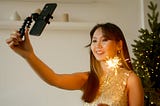An Asian woman with long dark hair and bangs holds a sparkler in one hand and her black cell phone on a selfie stick in the other. She is wearing a gold dress and smiling as she stands in front of a twinkling Christmas tree with white lights.