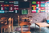 Mesas em um restaurante, com uma parede cheia de relógios ao fundo, cada relógio tendo a bandeira de um país.