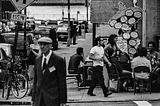 Black and white photo of a street corner in DUMBO, Brooklyn. An older man in a cap is crossing the street, while a woman behind him stands looking at her mobile device. People sit outside a Mexican restaurant as another pedestrian (or perhaps restaurant server) passes by.