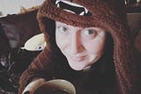 Selfie, a woman with long dark hair and blue eyes, sitting on the couch in her Chewbacca hoodie smiling and holding a hot drink in a mug.