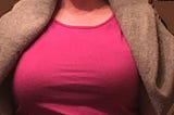 closeup of a very large chest straining the material of a pink shirt in multiple lines across the front and up and down in the shoulders to cover the collarbone; a large grey sweater drapes over the pale white shoulders.
