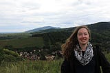 A tourist dressed in a wind jacket and floral scarf smiling in front of a backdrop of rolling hills, red-roofed houses and cloudy skies.