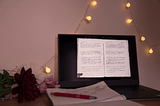 laptop with a State Archive document open, notebook with a family tree drawn on it, flowers on the bottom left corner and fairylights on the upper right corner; the picture’s rosy and warm colors remind of something from the past