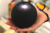Na imagem, uma mão segurando um Home Pod Mini da Apple, um dispositivo redondo de cor escura com uma luz brilhante no meio. Uma interface conversacional que permite com que as pessoas possam interagir com o ambiente e utilizar inteligências computacionais a partir de comandos de voz.