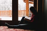 A woman reads a book on the carpet of her home with the natural light of a winter wonderland in her backyard illuminating the background.