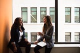 Two women in the workplace sitting on a windowsill and laughing