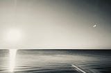 A symbolic monochromatic rendering of a photograph, with an almost invisible and forgotten sun, setting over the horizon of seemingly calm and undistrubed waters.