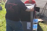 Two workers lifting an HVAC unit outside a house, preparing for installation near a window. One worker is wearing a cap and a dark T-shirt with the logo ‘Specialized Heating,’ while the other wears a red shirt and a baseball cap, focused on positioning the unit correctly. Green grass and autumn foliage can be seen in the background.