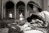 Gloria Swanson sits on a sun lounger in front of three arches in the façade of the house used for exterior shots in Sunset Boulevard. Her butler, played by Erich von Stroheim, hovers in the background