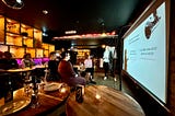 A female startup founder standing close to a pull-down projector screen, holding a microphone and practicing her pitch in front of 5–6 other startup founders sitting at small low club tables in a warm, softly orange glowing underground bar setting.