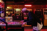 A boy in a white cap and black jacket leans over a pinball machine in a tattered arcade dimly lit by orange neon strips.