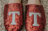 Feet wearing sparkly red slippers emblazoned with the letter T in silver