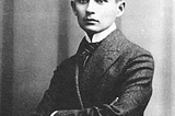 Mock-up of a young Franz Kafka photographed in b/w, looking soulful in a suit and tie, with a bright yellow hard hat superimposed on his head
