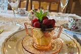 A table set with a lace tablecloth and floral dishes along with a pink glass teacup filled with fruit salad and garnished with fresh mint.