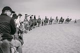 A group of people on camels follow a man into the desert.