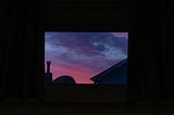 A photo of a pink and blue sunset through a window, only the sky is illuminated as the foreground is in shadow.