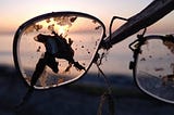 Cracked lens in an eye-glass frame held up by a stick with sun setting on the ocean in the background.