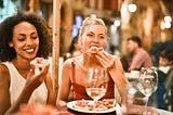 A blonde woman and a black woman sit eating pizza with wine glasses in front of them.