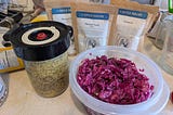 On the left, a glass jar with a fermentation lid (the lid has a valve in the center to release excess CO2 gas) containing yellow and brown mustard seeds and other ingredients. On the right, in a plastic container, reddish-purple sauerkraut made from red cabbage. In the background, three spice pouches from The Spice House — turmeric and two types of mustard seed.