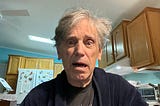 A surprised-looking man with disheveled gray hair wearing a dark robe and a T-shirt, standing in a kitchen. The background shows wooden cabinets, a refrigerator with colorful magnets, and a ceiling fan above. The expression on his face suggests shock or confusion.