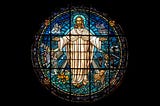 Stained glass image of Jesus with halo, dressed in white and arms open