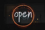 Neon “open” sign with white text, surrounded by orange ring, on a black background. Image has dim reflection.