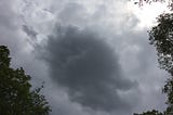 A dark cloud against lighter clouds, framed on three sides by tree branches.