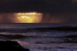 Dark raining clouds over swelling water, at the very horizon there’s a patch of sunshine, and a glimmer of hope.